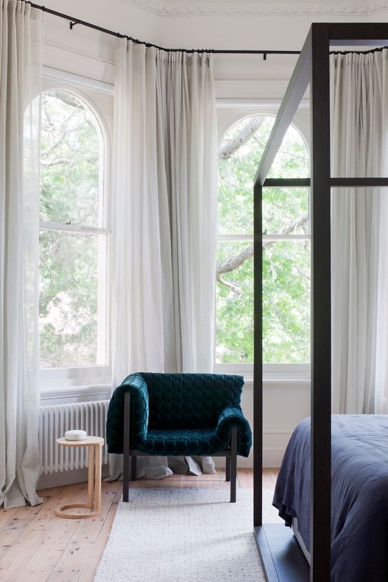 a sophisticated bedroom with arched double hung windows, a black canopy bed, a teal chair and a side table