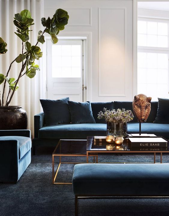 a sophisticated living room with navy velvet seating furniture, a dark grey rug, tiered glass coffee tables and greenery