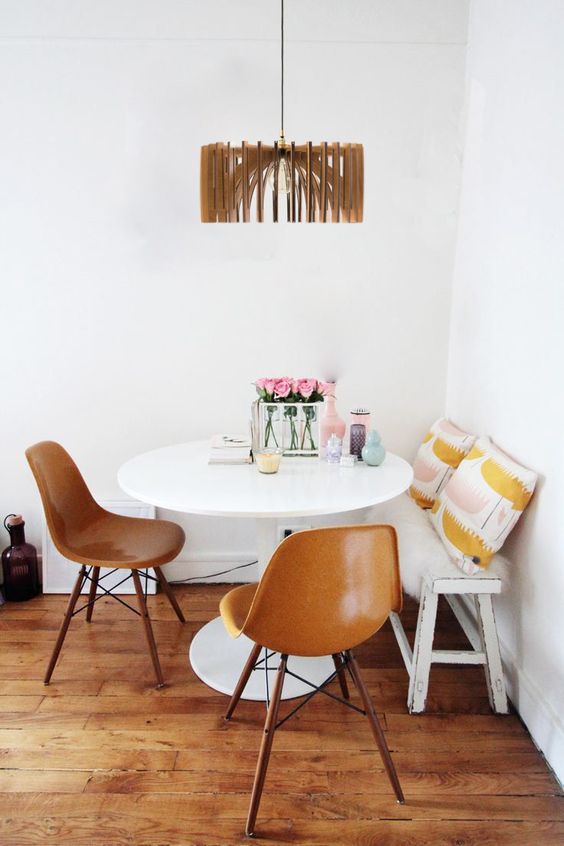 a stylish dining nook with a white bench and printed pillows, a round table, amber Eames chairs and a wooden lampshade