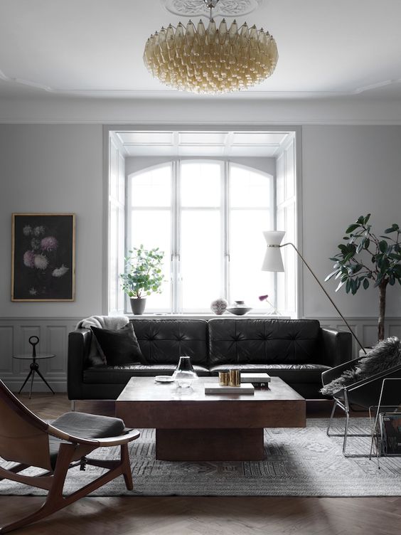 a stylish living room with a black leather sofa, a large coffee table, a stained plywood and metal chair, a gold chandelier and some art