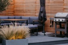 a stylish modern terrace with grain brushed basalt millboard decking, grey upholstered furniture, a wooden table, a concrete planter with lights and an umbrella