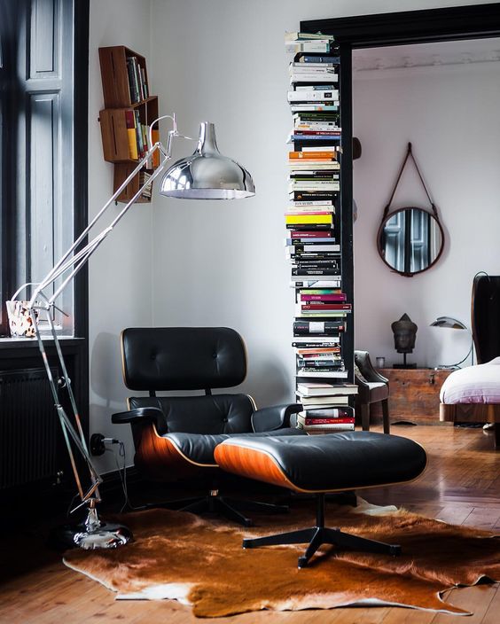 a stylish reading nook with a tall stack of books, a black Eames lounger and an ottoman, a metallic floor lamp and a rug