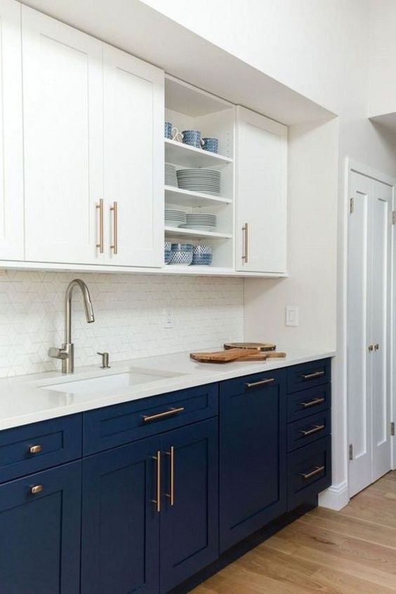 a stylish two tone kitchen with navy and white cabinets, shaker and open ones, white stone countertops, a white tile backsplash and brass fixtures