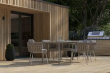 a terrace done with gold oak millboard decking, a metal dining table and woven upholstered chairs, potted plants and trees around