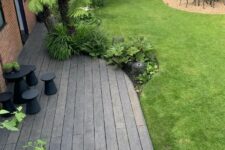 a tropical outdoor space with green grass, weathered oak decking, a black table and stools, trees and plants and a dining set