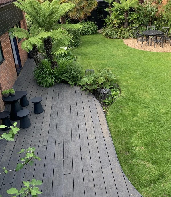 a tropical outdoor space with green grass, weathered oak decking, a black table and stools, trees and plants and a dining set