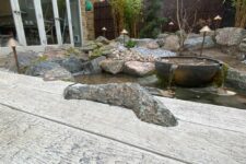 a weathered oak driftwood millboard deck with a stone, a natural pond next to it, rocks, pebbles and a bowl waterfall