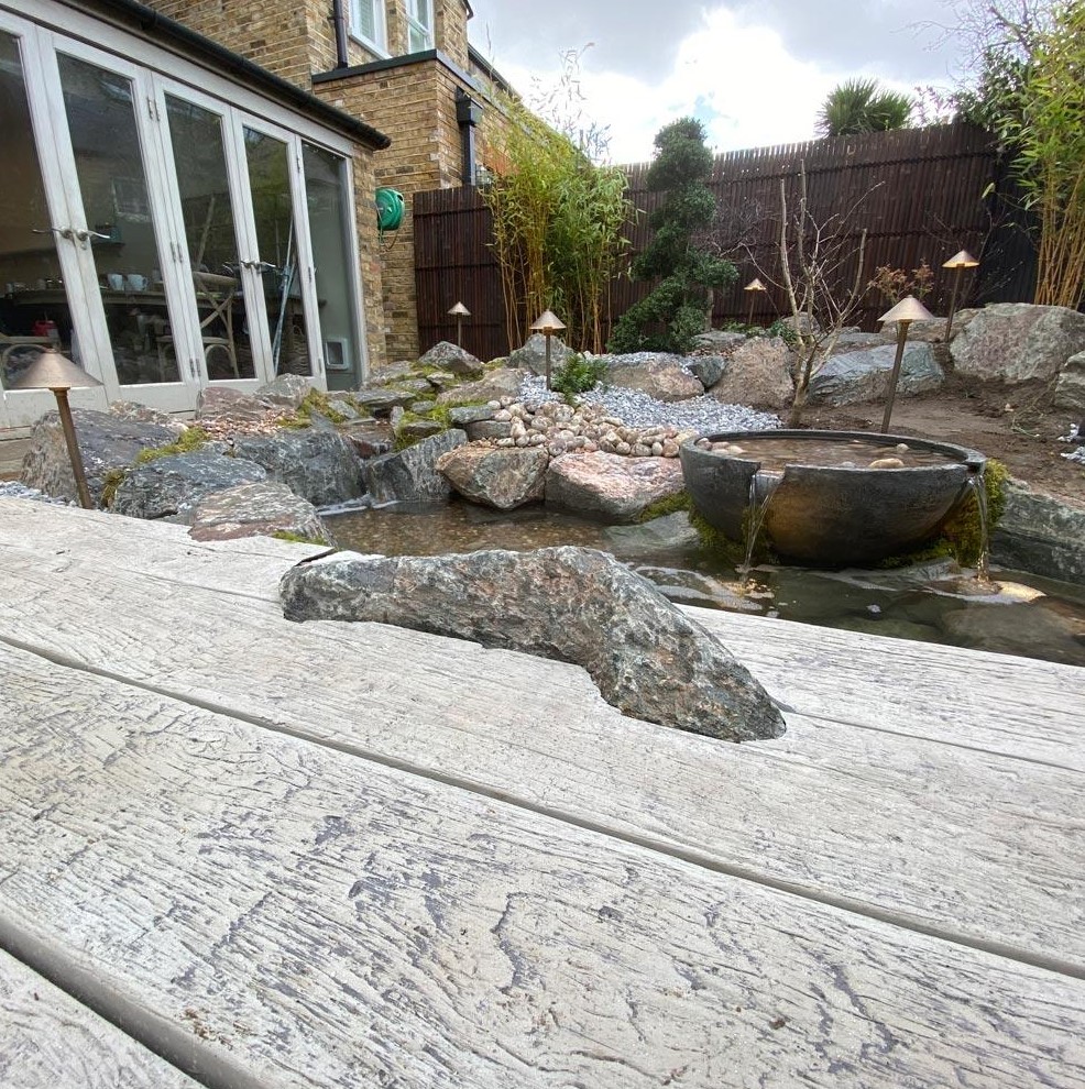 a weathered oak driftwood millboard deck with a stone, a natural pond next to it, rocks, pebbles and a bowl waterfall
