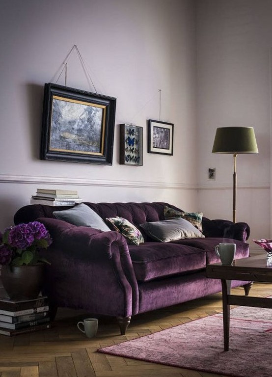 a welcoming living room with lilac walls, a refined purple velvet sofa, a lilac rug, a vintage gallery wall, a green floor lamp and a dark stained table