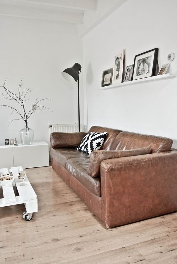 a white Scandinavian living room with a brown leather sofa, a low pallet coffee table on casters, a ledge gallery wall and a black floor lamp