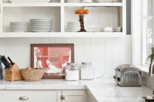 a white cottage kitchen with shaker cabinets and open upper ones, a white beadboard backsplash and white stone countertops