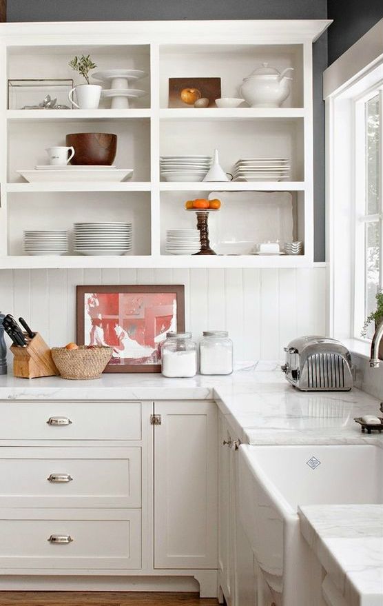 a white cottage kitchen with shaker cabinets and open upper ones, a white beadboard backsplash and white stone countertops