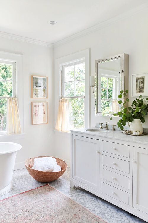 a white farmhouse bathroom with marble hex tiles, double hung windows, an oval tub, a large vanity and a window cabinet