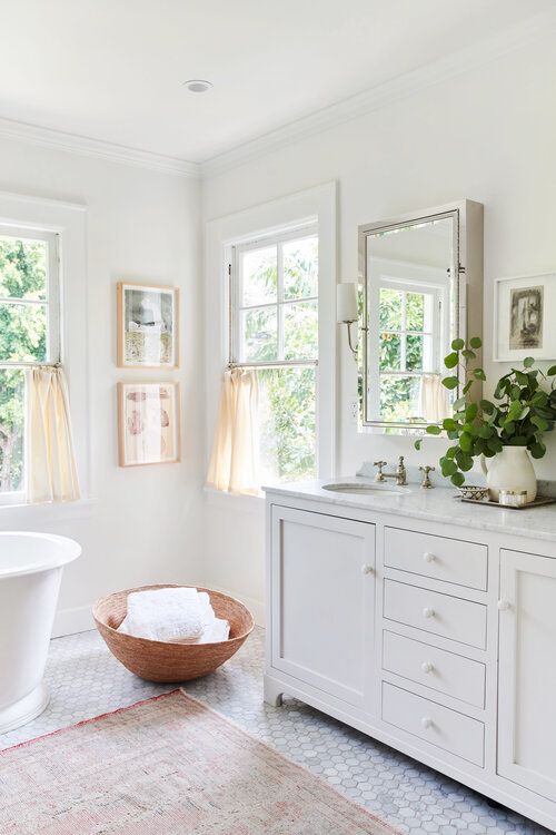 a white farmhouse bathroom with marble hex tiles, double-hung windows, an oval tub, a large vanity and a window cabinet