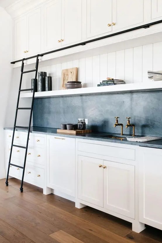 a white farmhouse kitchen with shaker cabinets, a black soapstone backsplash and countertops, open shelves and a black metal ladder