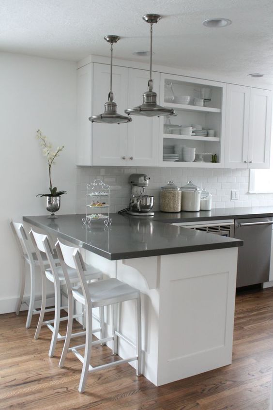 a white kitchen with shaker and open cabinets, black countertops, metal pendant lamps and a potted flower is cool and chic