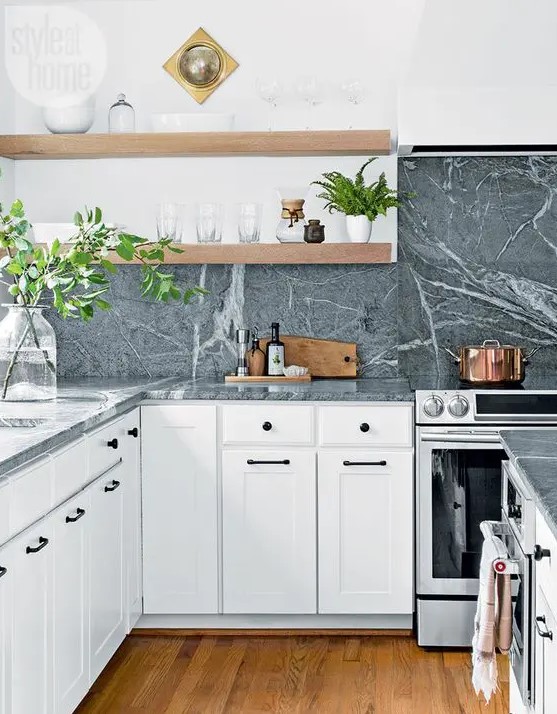 a white shaker style kitchen with open shelves, grey soapstone countertops and a backsplash, lots of greenery