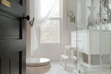 a white vintage bathroom with hex tiles and beadboard, a double-hung window, a large vanity and a glass-enclosed shower