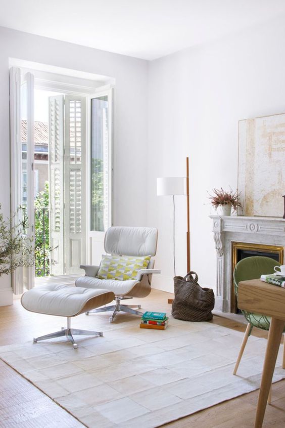 an airy white living room with a firpelace, a white Eames lounger and an ottoman, a floor lamp and a large artwork