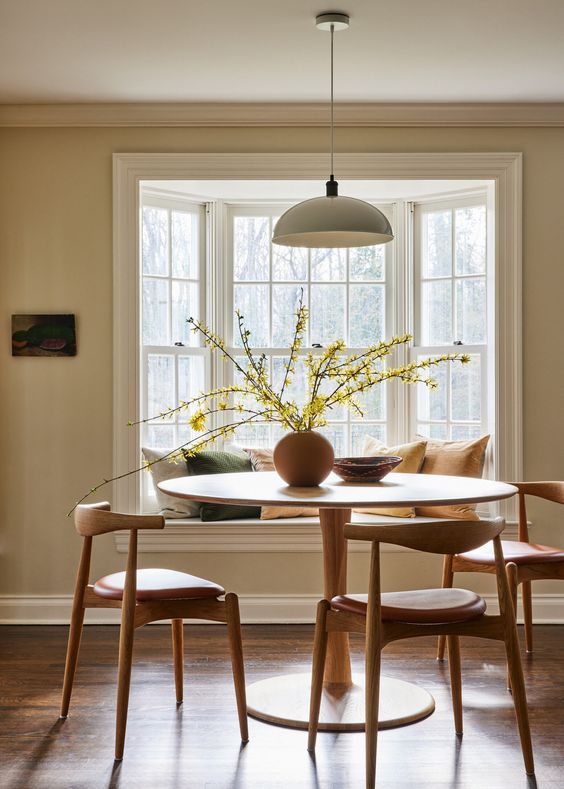 an earthy dining room with a bay window with a window seat, a round table and mid century modern chairs, a pendant lamp