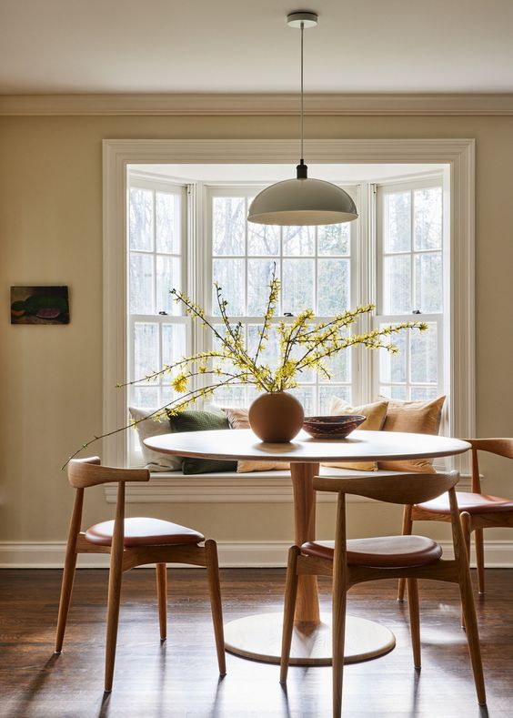 an earthy dining room with a bay window with a window seat, a round table and mid-century modern chairs, a pendant lamp