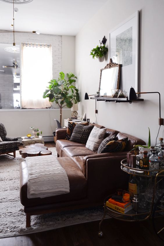 an eclectic living room with a brown leather sectional, a ledge with a mirror and art, some greenery and a home bar