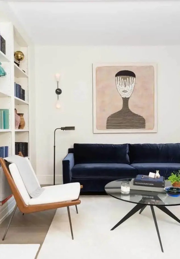 an elegant creamy living room with open storage units, a midnight blue velvet sofa, a chic chair, a bold artwork, a round coffee table and some greenery