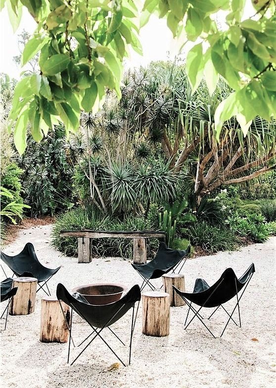 an outdoor fire pit space with a metal fire bowl, black leather butterfly chairs and tree stumps plus greenery around