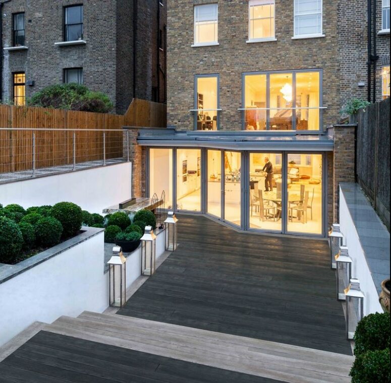 an outdoor space clad with charred and limed oak decking, with greenery in flower beds and lanterns that line up the pathway