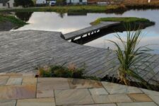 an outdoor space done with stone, with greenery growing, with a weathered oak millboard deck and a large pond is a very peaceful space