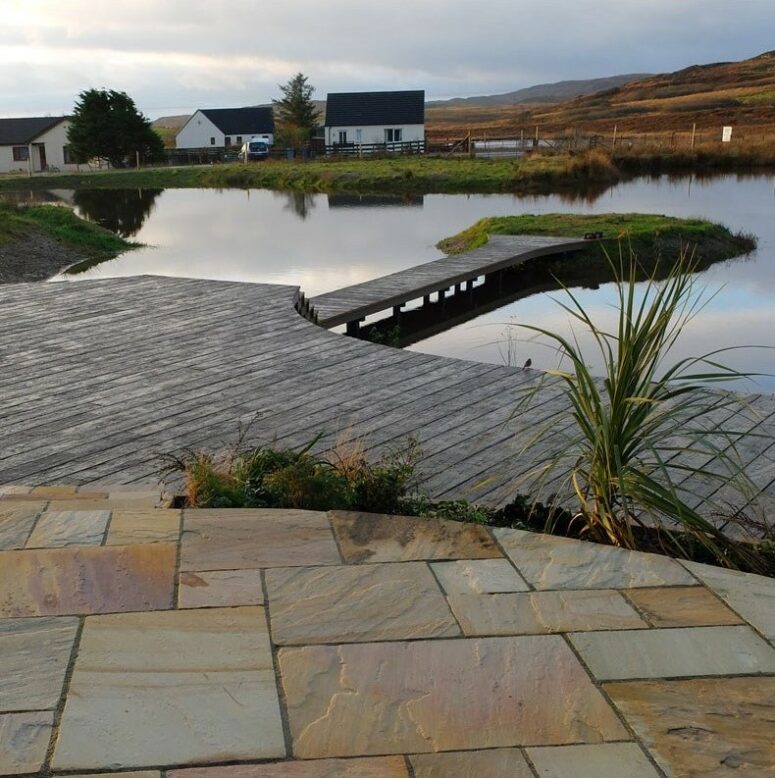 an outdoor space done with stone, with greenery growing, with a weathered oak millboard deck and a large pond is a very peaceful space