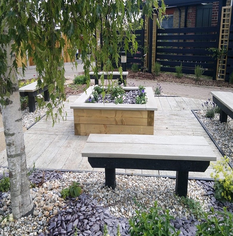 an outdoor space with weathered oak decking, benches, a flower bed with blooms, pebbles and trees