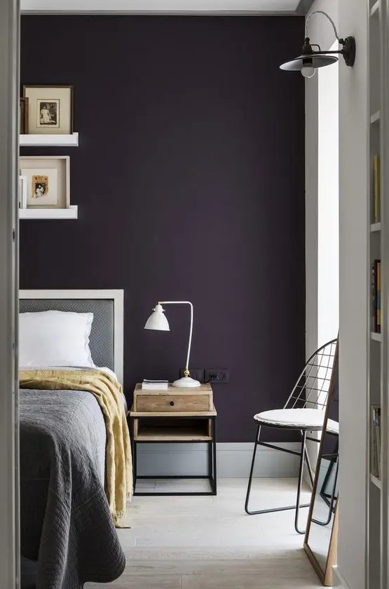 a stylish moody bedroom with a dark purple accent wall, grey and white furniture, a small nightstand with a table lamp and ledges with artworks