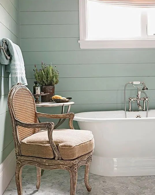 a mint green cottage bathroom with a vintage bathtub, a neutral antique chair, a side table with decor