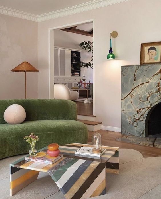 a welcoming living room with blush walls, a marble-clad fireplace, a grene curved sofa, a catchy striped sculptural coffee table