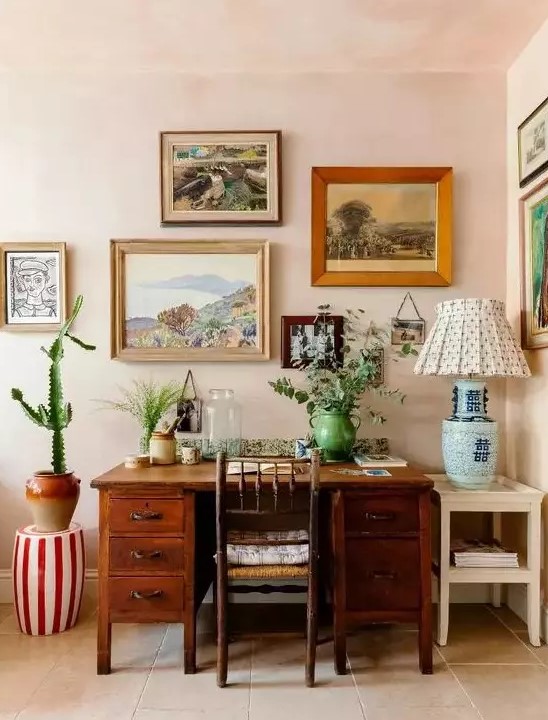 a bright mid-century modern home office with blush walls, a stained desk and a chair, a colorful gallery wall and greenery and a cactus