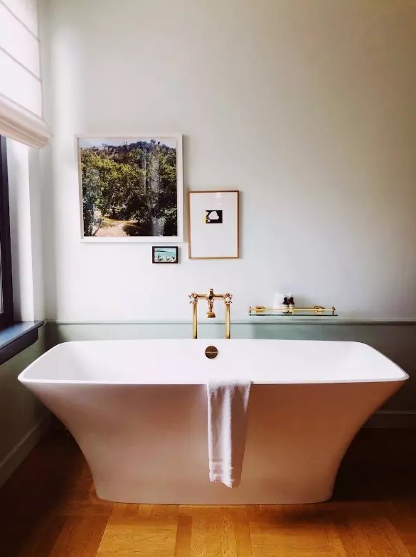 a chic and peaceful bathroom with green paneling, a catchily shaped bathtub, gold fixtures and a mini gallery wall with photos