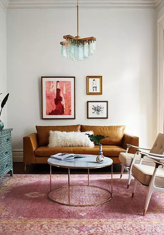 a colorful living room with a rust-colored leather sofa, beige chairs, a round table, a pink rug and a blue inlay dresser, a whimsical mini gallery wall