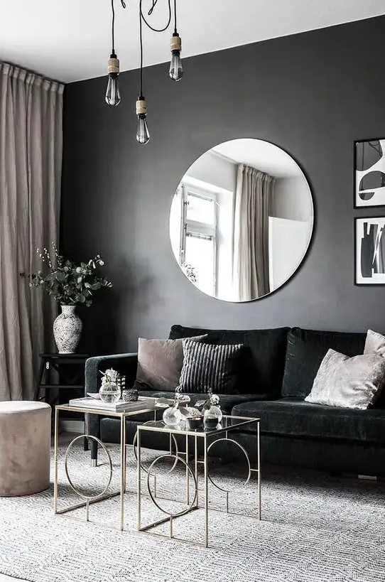 a chic and dramatic living room with a black accent wall, a black velvet sofa, chic glass coffee tables, pendant bulbs and a round mirror