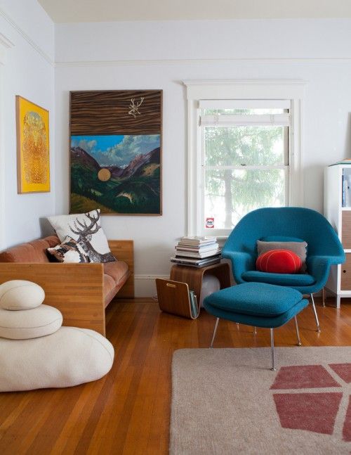 a bold mid-century modern living room with rust-colored loveseat, a blue Wumb chair with an ottoman, a curved shelf and pillows