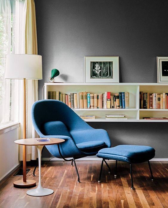 a bold mid-century modern reading space with a blue Wumb chair and ottoman, a side table and a bookshelf on the wall