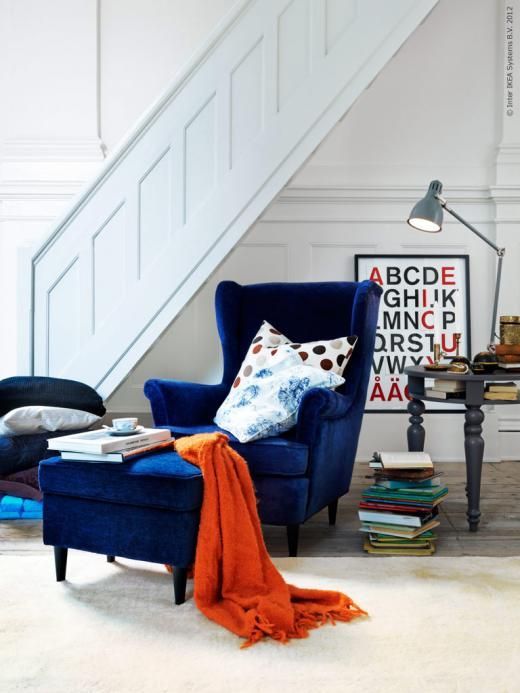 a bold reading nook with a navy Strandmon chair and an ottoman, a a grey table, book stacks and a table lamp is cool