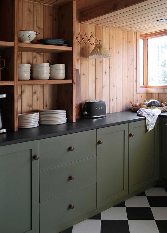 a cabin kitchen with stained wood walls, olive green lower cabinets, black stone countertops and a sconce