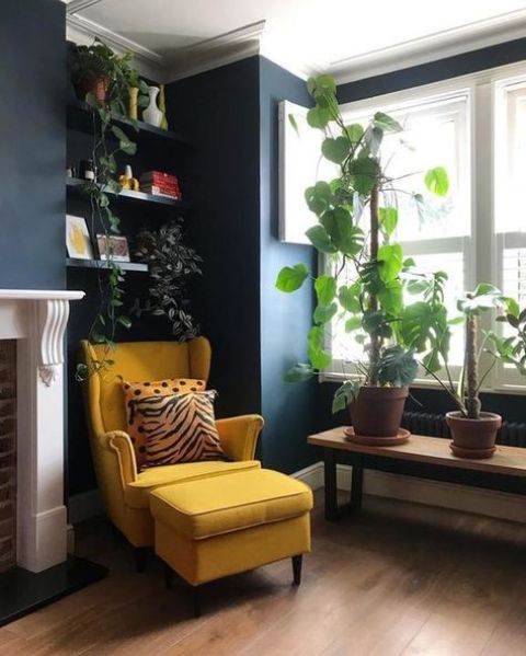 a cool nook by the window with navy walls, a bench with plants, built-in shelves with plants, a yellow Strandmon chair with an ottoman and a fireplace