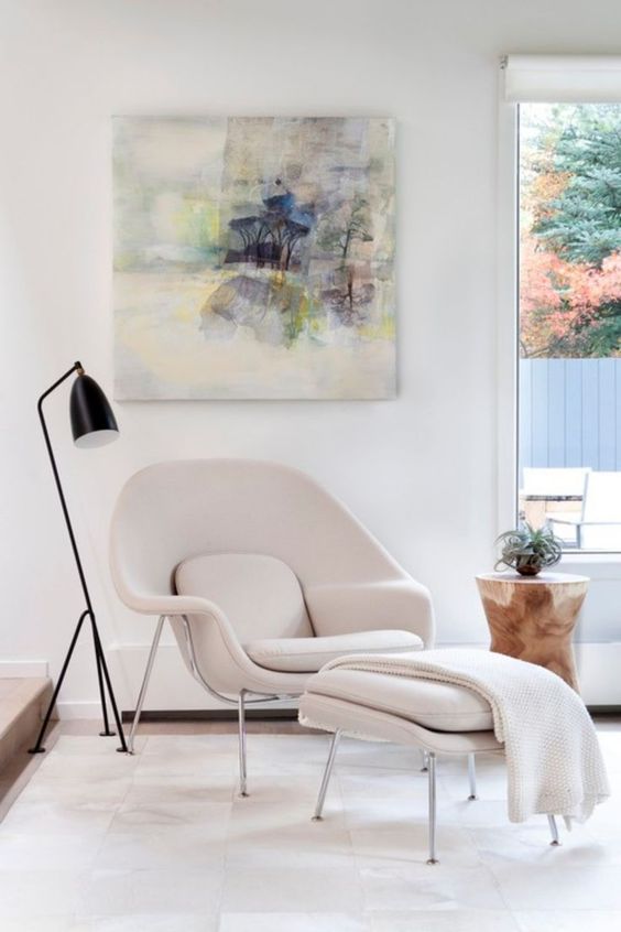 a delicate and airy space by the window with a creamy Wumb chair and ottoman, a black floor lamp and an artwork, a side table