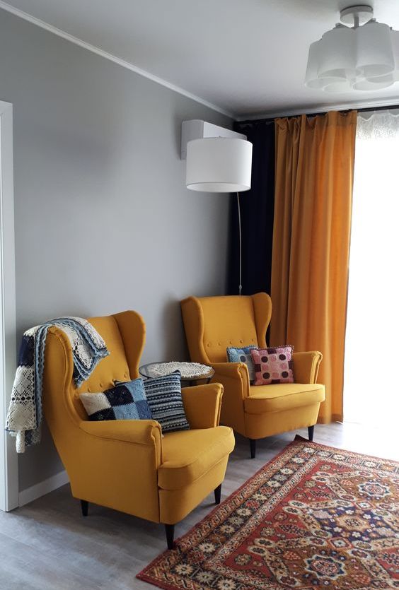 a mid-century modern living room with yellow Strandmon chairs, bright pillows and blankets, a bold printed rug and some lamps