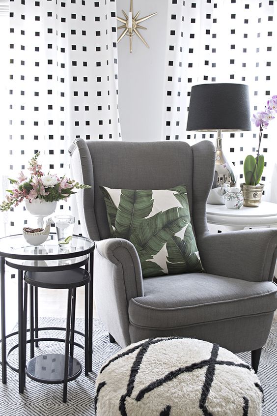 a modern cozy nook with a grey Strandmon chair, glass side tables, a printed pouf, a table lamp and printed curtains