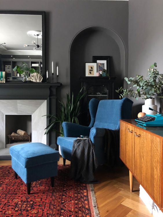 a moody living room grey walls, a fireplace, a mirror, a blue Strandmon chair and an ottoman, potted plants and a printed rug