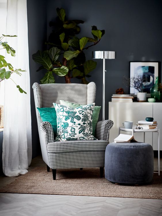 a moody nook with a graphite grey wall, a printed Strandmon chair, a grey pouf, a side table, a cabinet, potted greenery