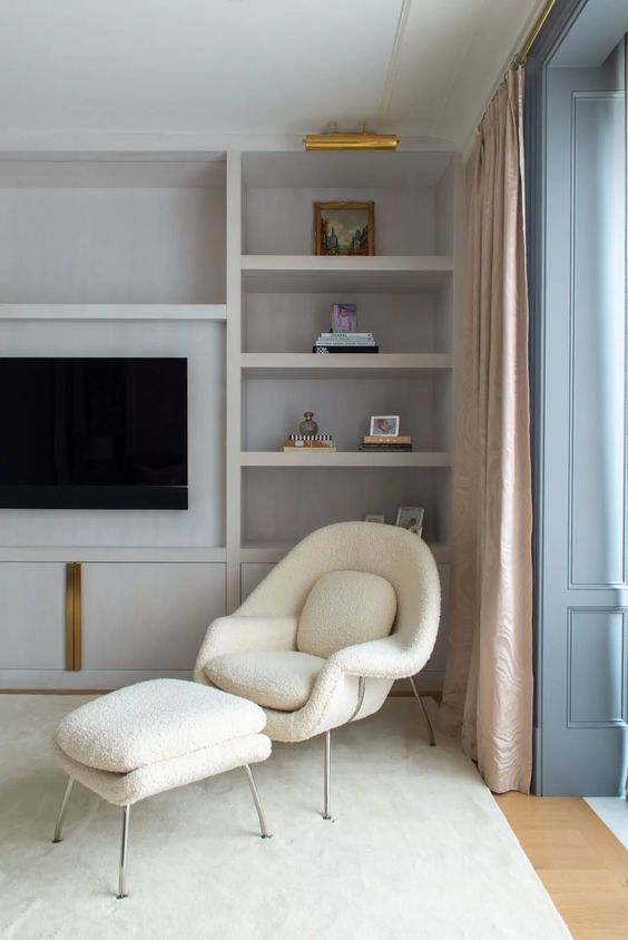 a peaceful neutral space with built-in shelves, a creamy Wumb chair and ottoman, neutral curtains and brass details
